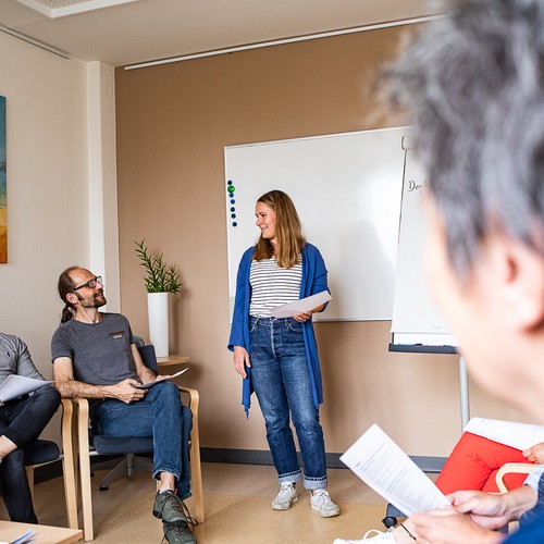 Eine Mitarbeiterin steht mit Unterlagen in der Hand vor einer Gruppe von Personen an einer Flipchart.