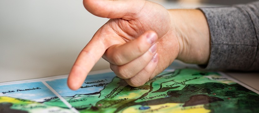 Eine Hand mit ausgestrecktem Zeigefinger zeigt auf einer gezeichneten Landkarte auf den Schriftzug "Gute Aussicht"